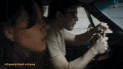 Movie gif. Swerving in a red Mustang, Aubrey Plaza as Sarah and Josh Hartnett as Danny in Operation Fortune: Ruse de Guerre frantically try to outrun a group of men shooting rifles from a black jeep.