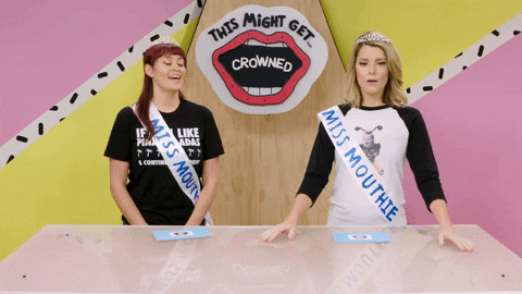 TV gif. Grace and Mamrie from This Might Get wear tiaras and sashes. Mamrie talks as Grace clasps her hands together and looks towards her. 