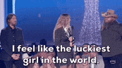 ACM Awards gif. Woman accepting award turns head around to speak into the microphone and says "I feel like the luckiest girl in the world" with a smile on her face while holding a trophy two male presenters stand behind her listening tentatively. 