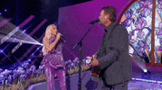 Academy of Country Music Awards gif. Gwen Stefani and Blake Shelton perform as a couple. The back of Shelton's faces the camera as strums the guitar strapped around his body and lovely looks at Stefani who maintains eye contact as she sings into a handheld mic with Blake. Gwen, wearing a sparkly purple outfit that matches the stage make a "C" shape with her hand and closes it like a crab claw in front of Shelton's face in a teasing manner.