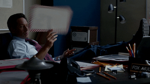 TV gif. David Duchovny as Mulder in the X-Files sits at a desk with his feet up, tossing a piece of mail over his shoulder.
