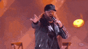 ACM Awards gif. Male musician walks across stage with flashing lights, nondescriptly speaks into mic with one hand and using the other to make an "i love you" hand signal. 