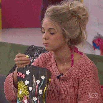 Reality TV gif. A woman with bed head is enthusiastically munching on popcorn. She reaches in the bag to grab more and turns to look at her friend.