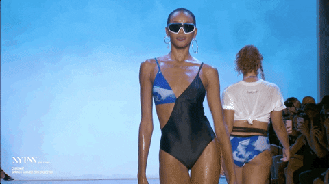 Video gif. Model struts down the runway in a swimming suit and dark sunglasses. She then waves her hand to fan herself like she’s too hot.