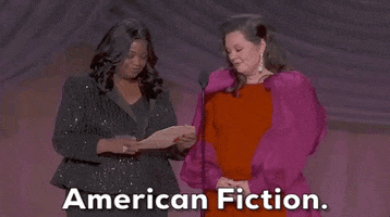 Oscars 2024 GIF. Melissa McCarthy and Octavia Spencer stand in front of the microphone. Spencer opens the envelope and announces the winner for Best Adapted Screenplay. She reads the envelope and looks up, saying, "American Fiction" and McCarthy reacts by sharply inhaling with excitement.