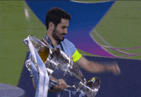 Sports gif. Rodri of Manchester City holds up the giant UEFA Champions League trophy and looks at us as he kisses and struts proudly across the field. 