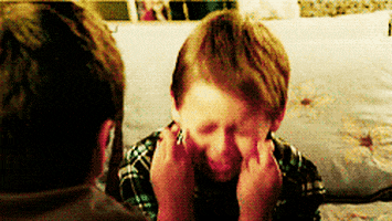 Video gif. Two little boys sit across from one another and one of them is closing his eyes tightly and crossing his fingers with all his might. He's hoping really, really hard for something to come true. 