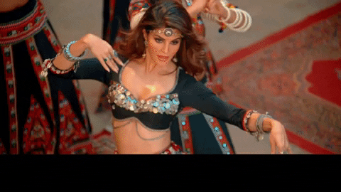 Video gif. A woman with long brown hair waves a ringed finger over her silver coined top. She glances up past a jeweled headband on her forehead. Women in colorful dresses and tops, dance behind her. Text, "Main panni panni ho gayi."