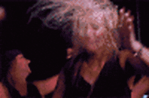 Video gif. Crowd of people at a rave with bright colorful lights flashing on them. A woman shakes her head with her mouth open, flings her arms around, and head bangs to the music.
