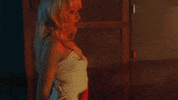 Music video gif from Sabrina Carpenter's music video for "Please Please Please." She wears a white dress and leans over Barry Keoghan, who is sitting in a chair with his hands behind his head. She dangles a pair of handcuffs in his face teasingly.