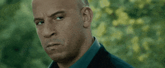 Movie gif. Vin Diesel as Dominic in Fast and Furious holds an intimidating gaze as a blurry green background whizzes by behind him.