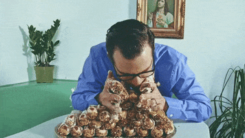 Music video gif. A scene from Weezer's music video where a man is gorging on a mountain of cannolis. He sticks his entire face in the cannolis, eating palmfuls of the desert, but eventually ends up lifting the entire plate to his mouth and leaning back, giving himself a cannoli facial. 