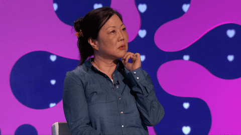 Reality TV gif. Margaret Cho on The Celebrity Dating Game. She's sitting in a chair pondering with her hand on her chin before coming to a realization and saying, "Oh," and looking at us suddenly.