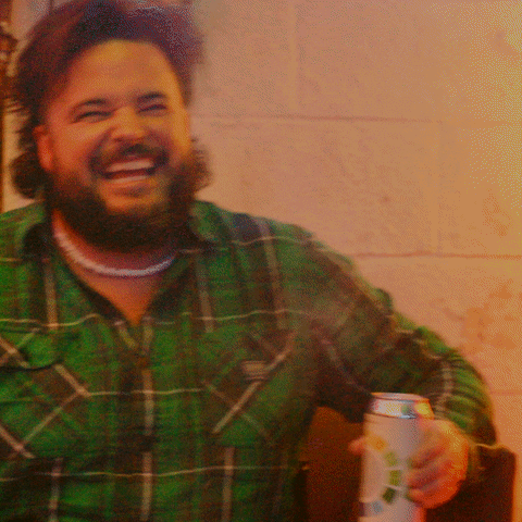 Celebrity gif. Jon Gabrus holds a beer as he sits on a couch and cracks up laughing as colorful lights flash around him