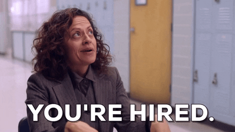 A woman is shaking someone's hand and saying the words, "YOU'RE HIRED."