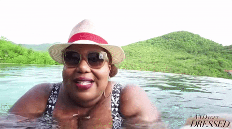 Video gif. From her Youtube channel And I Get Dressed, Kellie Brown wears a straw hat and sunglasses happily swimming in calm water at the beach with a background of lush greenery. 