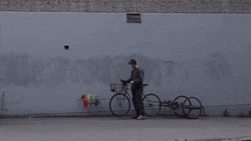 Video gif. Device attached to a bike has a long rod that paints a perfect rainbow arc on a gray concrete wall. The bike rider watches the entire motion as the paint travels smoothly over his head.