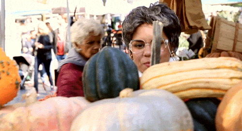 Video gif. Woman dressed as a grandmother wears a neck brace as she scans squash at a farmer's market. She smiles with delight until she lays eyes on one with warts and scrunches her nose in disgust.