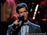 Video gif. A man in a sport jacket stands on stage, sighs heavily and mutters into a microphone.
