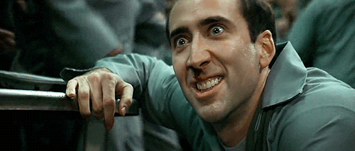 Movie gif. Wearing a blue jumpsuit and leaning against the edge of a metal table, Nicolas Cage seems to be growling at us while a chaotic scene unfolds in the background.