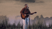 TV gif.  Noah Kahan performing on stage at the 2024 ACM Awards. Noah sings into a microphone, wearing a brown jacket and playing an acoustic guitar. The background features a scenic backdrop with soft lighting.