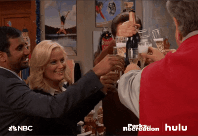 Parks and Recreation gif. Amy Poehler, Aziz Ansari, Nick Offerman, and Jim O'Heir as Leslie, Tom, Ron, and Jerry cheerfully clink champagne glasses with each other and other colleagues.