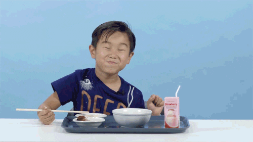 Video gif. Boy shimmies side to side as he chews and smiles with his eyes closed while holding a chopstick over a tray of food.