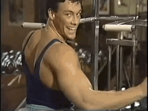 Celebrity gif. Jean Claude Van Damme does the center splits over a weight set as he winks over his muscular shoulder and gives a wave. 