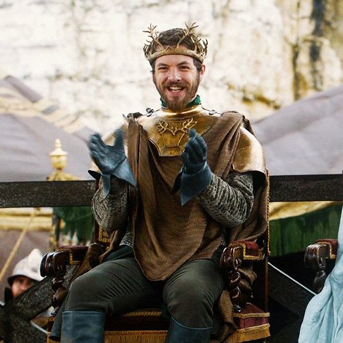 Game of Thrones gif. Gethin Anthony as Renly Baratheon applauds from a high chair, laughing and then winking subtly.
