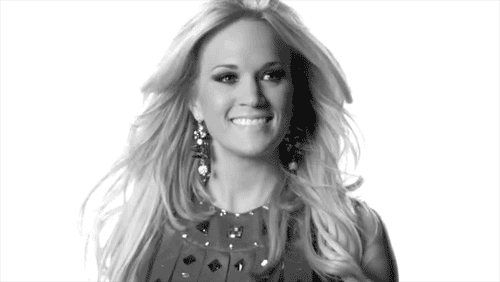 Celebrity gif. Carrie Underwood smiles with a shrug, her hair blowing slightly.
