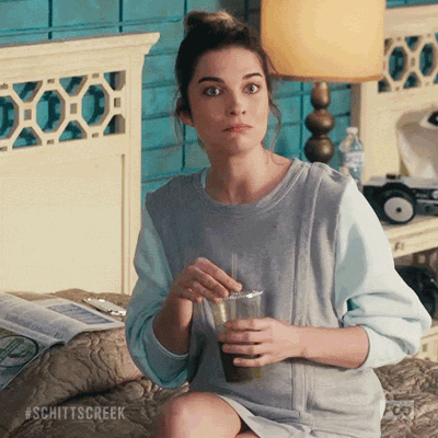 Schitt's Creek gif. Annie Murphy as Alexis Rose sits on a bed holding an iced coffee as she gives us a confused, wide-eyed shrug.