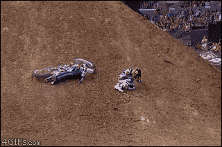 Sports gif. Dirt track racer sits on the track, wincing in pain, with his bike laying next to him. Two men run onto the field to help the racer. One goes up to help him get up and another picked up the dirt bike. When the man picks up the dirt bike, he loses control of it and it slams right into the racer’s back, causing him to fly back onto the ground.