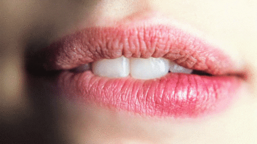 Video gif. Close-up on a woman's pink lips as she bites the side of her bottom lip in a sexy, sultry manner. 