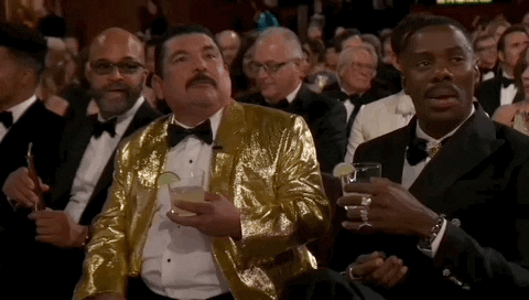 Oscars 2024 GIF. Guillermo sits next to Colman Domingo in the crowd and both are holding margaritas. Colman has his legs crossed and he waves a hand out increduously, as if it should be a given that he's going to be drinking a margarita. 