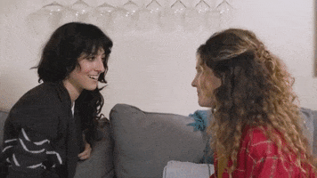 TV gif. Two women sit on opposite ends of a couch while staring and smiling at each other, before one goes in for a kiss. They kiss passionately, removing their jackets and embracing each other.