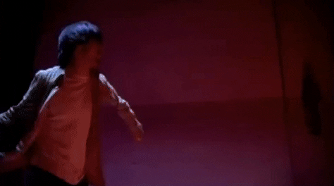 Music video gif. Dave Grohl in the "Walking After You" music video throws a chair across a room with high ceilings cast in a red light as a spotlight shines on him. 