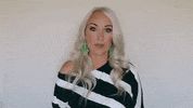 Video gif. A woman looks straight at us with a blank expression and then looks up, lifts her hands up to point above her. She looks back at us and nods with a smirk on her face. 