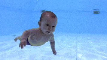 Video gif. A baby waves his arms and kicks his legs as he swims underwater in a pool.