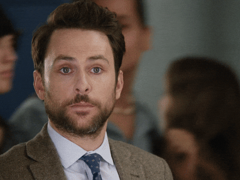 Movie gif. Wearing a brown suit jacket, Charlie Day gives us a blank look as several people walk by him in the background. After a second, he raises a hand to give us a thumbs up, then walks out of frame.
