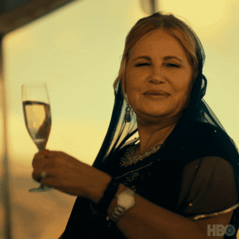 TV gif. Jennifer Coolidge as Tanya McQuoid in "The White Lotus" is wearing a gold-speckled black chiffon veil over her head on a swaying boat. She awkwardly cracks a smile as she raises a glass of champagne and says "Wee-hee!" 