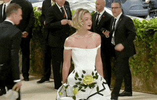Met Gala 2024 gif. Gigi Hadid gives a calm smile for photos wearing a cream colored Thom Browne dress that is fitted through the drop waist with off-the-shoulder straps that fit tightly around her upper arm. The dress flares at her hips with green leaves and pale yellow roses.