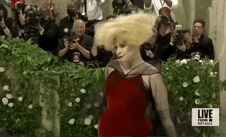 Met Gala 2024 gif. Gwendoline Christie wearing a red Maison Margiela gown and bold, oversized blonde wig, fluffs her sheer black cape, maintaining her posture as she prepares to pose.