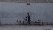 Video gif. Device attached to a bike has a long rod that paints a perfect rainbow arc on a gray concrete wall. The bike rider watches the entire motion as the paint travels smoothly over his head.