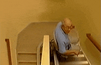 Video gif. An old man rides a chair lift assist down the stairs.