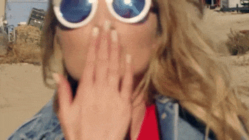 Music video gif. From the video for Cheat Codes' Feels Great, a blonde-haired woman wearing sunglasses blows a kiss up-close at us.