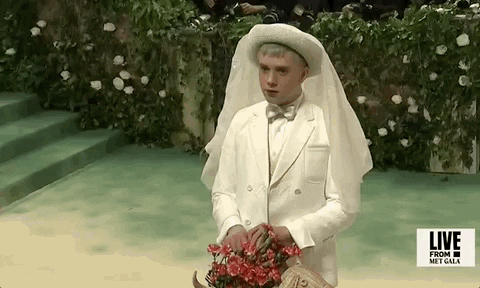 Met Gala 2024 gif. Cole Escola wears a Thom Browne monotone ivory-colored jacket, collared shirt, and bowtie holding a tan wicker dog-shaped basket with pink carnations emerging from the top. They are wearing a circular brimmed hat with a sheer ivory veil hanging down the back. 