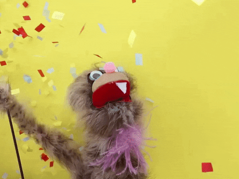 Video gif. Light-brown furry puppet with big red open mouth and one sharp tooth waves arm wildly as colorful confetti pours down from above.