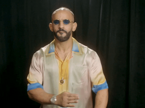Video gif. Ottman Azaitar wearing a silk shirt and blue tinted glasses strikes a karate pose in front of a black curtain and says, "Come at me bro."
