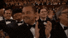 Celebrity gif. Leonardo DiCaprio sits in the audience of the Academy Awards. He looks over his shoulder with a large smile and claps along with the crowd. 