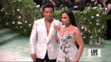 Met Gala 2024 gif. Demi Lovato poses for the cameras with Pabral Gurung, wearing his smokey lavender gown embellished with silver flowers, as he waves and adjusts.